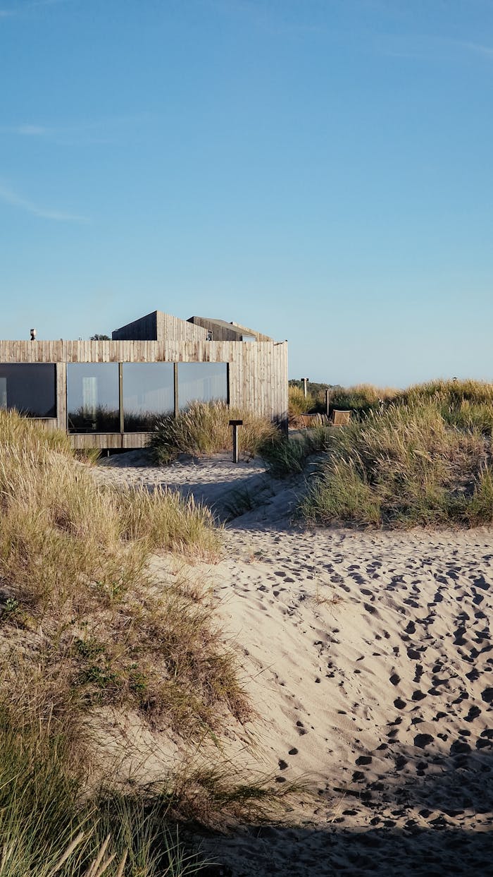 A modern wooden house sits peacefully amidst sandy dunes under clear skies.
